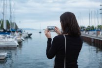 Вид сзади женщины, щёлкающей фото с мобильного телефона возле гавани — стоковое фото