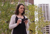 Жінка має каву під час використання мобільного телефону в місті — стокове фото