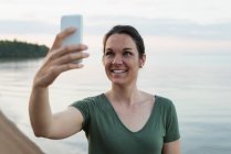 Mujer feliz tomando selfie en el teléfono móvil cerca de riverside - foto de stock