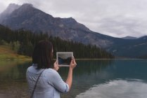 Vista trasera de la mujer haciendo clic fotos con la tableta digital cerca de la orilla del lago - foto de stock