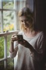 Красива жінка стоїть біля вікна і використовує мобільний телефон вдома — стокове фото