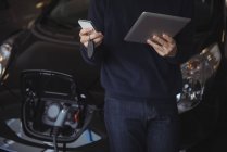 Середина людини, яка використовує цифровий планшет і мобільний телефон, заряджаючи електромобіль в гаражі — стокове фото