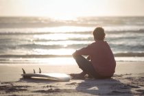 Mann mit Surfbrett sitzt in der Abenddämmerung am Strand — Stockfoto