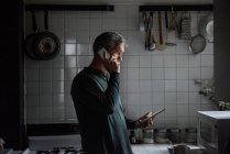 Мужчина разговаривает по телефону и использует планшет на кухне — стоковое фото