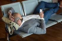 Uomo d'affari che utilizza il telefono cellulare mentre è sdraiato su sedie in sala d'attesa presso il terminal dell'aeroporto — Foto stock