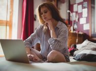 Задумчивая женщина использует ноутбук в спальне дома — стоковое фото