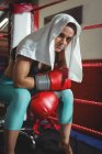 Уставшая боксерша с полотенцем сидит в ринге в фитнес-студии — стоковое фото