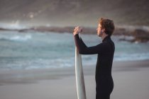 Vista lateral de um homem de fato de mergulho em pé na praia com prancha de surf — Fotografia de Stock