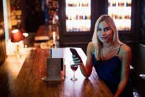 Retrato de mulher usando telefone celular com vinho tinto na mesa no bar — Fotografia de Stock