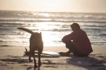 Mann mit Surfbrett sitzt in der Abenddämmerung mit Hund am Strand — Stockfoto