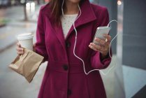 Businesswoman holding disposable coffee cup and parcel while listening to music near office building — Stock Photo