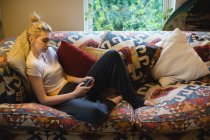 Femme couchée et utilisant un téléphone portable sur le canapé dans le salon — Photo de stock