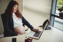 Беременная деловая женщина трогает живот во время использования ноутбука в офисе — стоковое фото