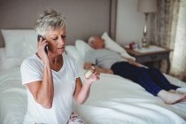 Беспокойная пожилая женщина сидит в спальне с лекарствами и разговаривает по мобильному телефону — стоковое фото