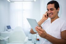 Médico sosteniendo tableta digital mientras habla en el teléfono móvil en la clínica - foto de stock