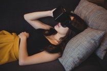 Frau benutzt Virtual-Reality-Headset, während sie zu Hause auf dem Sofa liegt — Stockfoto