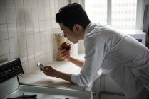 Человек, использующий мобильный телефон, имея кекс дома — стоковое фото