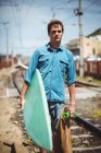 Porträt eines Mannes, der Skateboard und Surfbrett über Bahngleise trägt — Stockfoto
