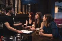 Amigos sosteniendo vasos de cerveza en el mostrador del bar e interactuando con el camarero - foto de stock