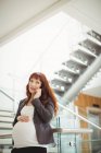 Беременная деловая женщина разговаривает по мобильному телефону у лестницы в офисе — стоковое фото