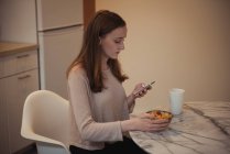 Mujer usando el teléfono móvil mientras desayuna en la cocina en casa - foto de stock