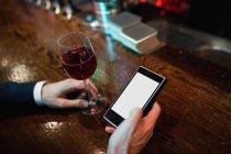 Mains d'homme d'affaires utilisant un téléphone portable avec un verre de vin rouge à la main au bar — Photo de stock