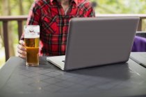 Seção média do homem usando laptop com copo de cerveja na mesa no bar — Fotografia de Stock