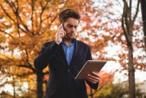Businessman talking on mobile phone and holding digital tablet in autumn — Stock Photo