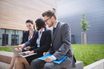 Businesswoman sitting with colleagues and using laptop outside office building — Stock Photo