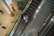 Empresários interagindo uns com os outros enquanto descem na escada rolante no terminal do aeroporto — Fotografia de Stock