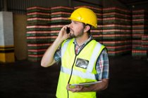 Male worker holding clipboard while talking on phone in factory — Stock Photo