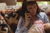 Мати розмовляє на мобільному телефоні під час грудного вигодовування дитини на дивані у вітальні — стокове фото