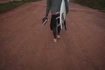 Низкая часть человека с доской для серфинга и обувью, идущего по дороге — стоковое фото