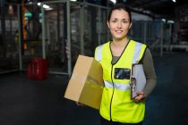Retrato de una joven trabajadora cargando caja y portapapeles en almacén - foto de stock