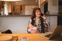 Mother carrying baby while talking on mobile phone at home — Stock Photo