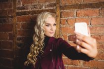 Hermosa rubia de pie contra la pared de ladrillo y tomando una selfie en su teléfono móvil - foto de stock