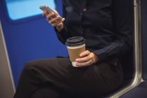Sección media de la mujer de negocios con taza de café usando el teléfono mientras está sentado en el tren - foto de stock