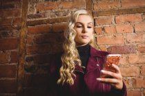 Schöne blonde Frau, die mit Handy gegen Ziegelmauer steht — Stockfoto