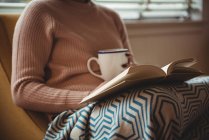 Mulher lendo livro enquanto toma café na sala de estar em casa — Fotografia de Stock