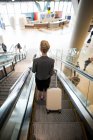 Vista posteriore della donna d'affari in piedi sulla scala mobile con bagagli in aeroporto — Foto stock