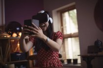 Счастливая женщина использует гарнитуру виртуальной реальности, держа дома мобильный телефон — стоковое фото