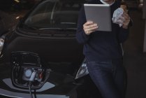 Man using digital tablet and snacking while charging electric car in garage — Stock Photo