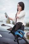 Красивая женщина разговаривает по мобильному телефону во время зарядки электромобиля на улице — стоковое фото