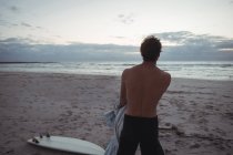 Rear view man standing on beach with surfboard at dusk — Stock Photo
