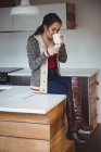 Жінка п'є чашку кави, використовуючи мобільний телефон на кухні вдома — стокове фото