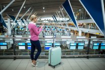 Female commuter with luggage using mobile phone in airport — Stock Photo
