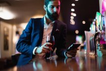 Empresário usando telefone celular com copo de vinho tinto na mão no bar — Fotografia de Stock