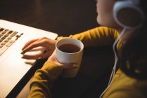 Женщина пьет кофе во время работы на ноутбуке в кабинете дома — стоковое фото