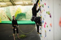Trainer assisting man while climbing on artificial wall in gym — Stock Photo