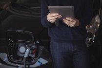 Partie médiane de l'homme en utilisant une tablette numérique tout en rechargeant la voiture électrique dans le garage — Photo de stock